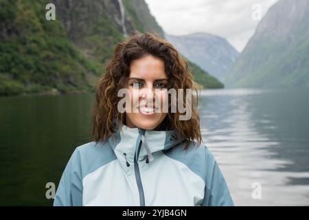 Nahaufnahme Porträt einer jungen brünetten Frau im Alter von 30 Jahren mit Regenjacke, die in die Kamera blickt, mit einer üppigen grünen Berglandschaft und einem See Stockfoto
