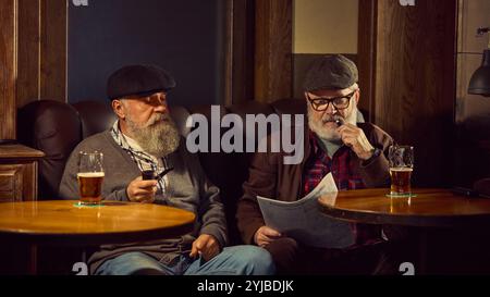 Zwei ältere, bärtige Männer in Mützen sitzen in Ledersesseln, rauchen Tabakpfeifen, lesen Zeitung und trinken Biene in der gemütlichen Bar. Stockfoto