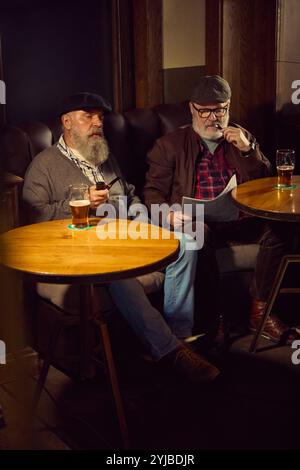 Ein älterer bärtiger Mann sitzt in einer Bar, einer liest Zeitung, der andere raucht Pfeife und genießt einen ruhigen und entspannenden Moment. Stockfoto