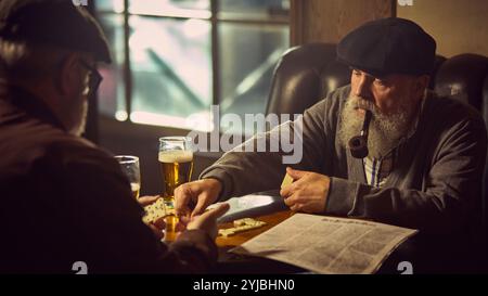 Älterer Mann mit Pfeife im Mund, der mit seinem Freund Dominosteine in warmer, gemütlicher Bar spielt, Bier trinkt, ruhiger Freizeit-Moment. Stockfoto