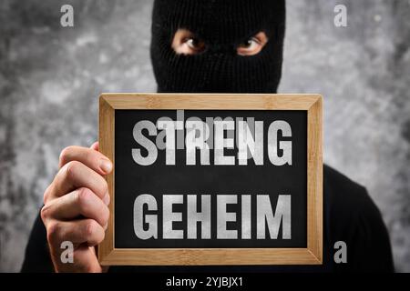 Bayern, Deutschland - 14. November 2024: Streng geheim, sagt ein Schild vor einer maskierten anonymen/unbekannten Person. Symbolisches Bild für geheime und vertrauliche Informationen, die nicht veröffentlicht werden sollten. FOTOMONTAGE *** Streng geheim, stehen auf einem Schild vor maskierter anonymer / unbekannter Person. Symbolbild für geheime und vertrauliche Informationen die nicht an die Öffentlichkeit kommen sollen. FOTOMONTAGE Stockfoto