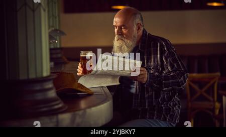 Älterer bärtiger Mann in kariertem Hemd, der an der Bar mit warmem Licht sitzt, Bier trinkt und Zeitung liest. Entspannende Atmosphäre Stockfoto