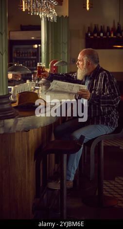 Älterer bärtiger Mann in kariertem Hemd, der an der Bar mit warmem Licht sitzt, Bier trinkt und Zeitung liest. Entspannende Atmosphäre Stockfoto