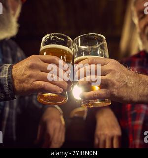 Zwei ältere Männer, die Gläser mit schäumendem Bier anklitzen. Konzentrieren Sie sich auf Hände und Brillen. Warm beleuchtete Leiste. Stockfoto