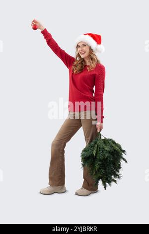 Frohe Weihnachten, glückliche lächelnde Frau, die Baumspitze und roten Ball in der Hand hält, um den Weihnachtsbaum zu machen und zu dekorieren, und in die Kamera schaut, mit Weihnachtsmütze Stockfoto