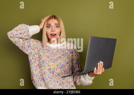 Eine überraschte junge Frau in einem farbenfrohen Strickpullover hält einen Laptop vor einem grünen Hintergrund und drückt so Schock und Staunen aus. Stockfoto