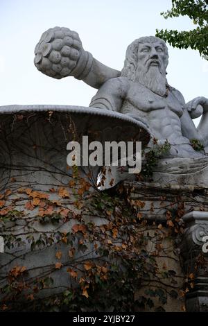 Eine Steinstatue eines muskulösen bärtigen Mannes, der einen Schläger hält, mit Herbstelfeu, der die Basis hochklettert. Alter, mythologischer Look. Stockfoto