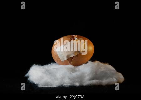 Gebrochene Eierschale mit Reparaturpflaster auf einer Wattwolke. Das Konzept der Fragilität und Resilienz Stockfoto