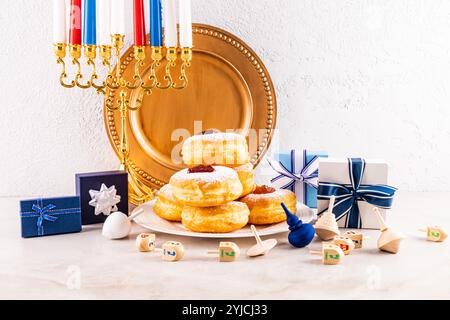 Gebratener Donut mit Puderzucker und Marmelade auf einem Teller, Menora, Dreidels, die Schachteln mit Geschenken. Strukturierter Wandhintergrund. Das Konzept der Hanukka Stockfoto