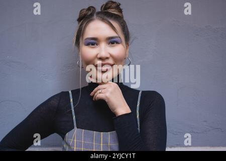 Stilvolle asiatische Frau mit modernem Make-up und Space Buns-Frisur, die vor minimalistischem grauem Hintergrund posiert Stockfoto