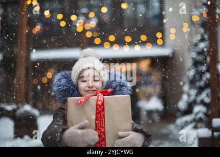 Porträt eines fröhlichen Mädchens mit einer Geschenkbox zu Weihnachten auf einer Stadtstraße im Winter mit Schnee auf einem festlichen Markt mit Dekorationen und Feenlichtern. Warm Stockfoto