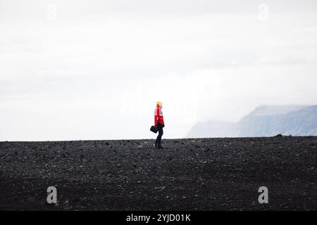 Ein Reisender in roter Jacke, der an einem schwarzen Sandstrand in Island steht. Nur eine Person, kaltes, nebeliges isländisches Wetter Stockfoto