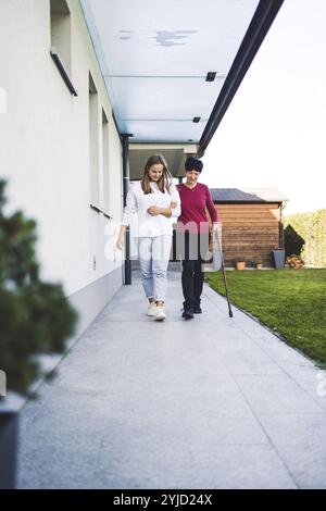 Junge kaukasische Frau, Krankenschwester, bei einem Hausbesuch, um eine Seniorin zu versorgen. Die Schwester hilft einer Seniorin bei den täglichen Aufgaben, hilft ihr, zu laufen Stockfoto