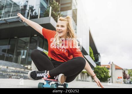 Junge blonde weiße Mädchen, die draußen Spaß auf einem Skateboard hat, schwarze Leggings und rotes Hemd, Sonnenlicht. Teenager lacht, hat draußen Spaß Stockfoto