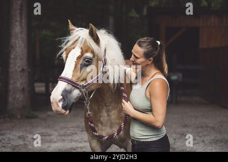 Kaukasische Frau kümmert sich um ihr Pferd im Stall und draußen, putzt sein Har mit einer Haarbürste. Wunderschönes, freundliches braunes Pferd, das geduldig wartet Stockfoto