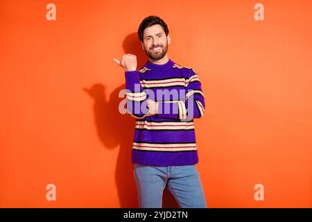 Lächelnder junger Mann in gestreiftem Pullover, der auf orangefarbenen Hintergrund zeigt und Glück und lässigen Fashion-Stil zeigt Stockfoto