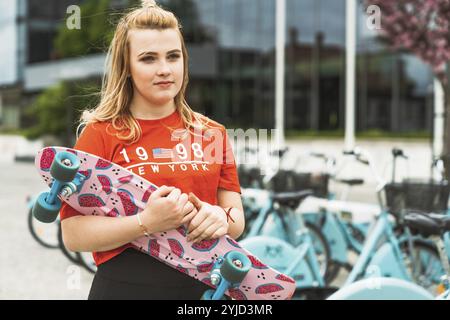 Junge blonde weiße Mädchen, die draußen Spaß auf einem Skateboard hat, schwarze Leggings und rotes Hemd, Sonnenlicht. Teenager lacht, hat draußen Spaß Stockfoto