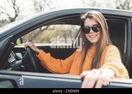 Schöne junge, glückliche, lächelnde weiße Frau, die in ihrem Auto fährt, Sonnenbrille und einen orangefarbenen Pullover trägt. Stockfoto