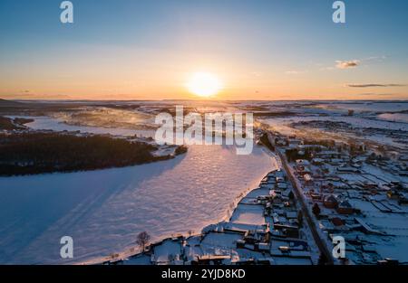 Sonnenaufgang über schneebedeckter Landschaft, der einen Fluss und ein ruhiges Dorf im Winter auf dem Land offenbart Stockfoto