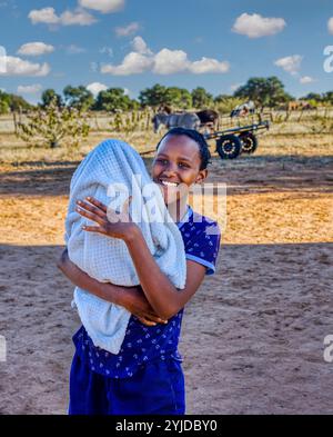 afrikanisches Dorfleben, junges Mädchen, das ein Baby hält, geflochtene Haare trägt, Eselkarre im Hintergrund Stockfoto