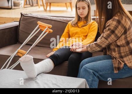 Ein Mädchen mit gebrochenem Bein sitzt mit Mutter zu Hause auf der Couch. Trauriges kleines Kind, das Teddybären umarmt und sich von gebrochenem Bein erholt. Mädchen mit Gusskrücken für schnelle Rehabilitation. Mutter, die Tochter unterstützt Stockfoto