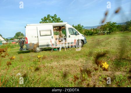 Ein Camper steht auf einer Wiese eines Bauernhofs. Eine Frau sitzt am gedeckten Tisch auf einem Stuhl vor dem Camper. Rheinland-Pfalz, Deutschland. Mit einem Campervan unterwegs *** Ein Wohnmobil steht auf einer Wiese auf einem Bauernhof Eine Frau sitzt an einem gedeckten Tisch auf einem Stuhl vor dem Wohnmobil Rheinland-Pfalz Stockfoto