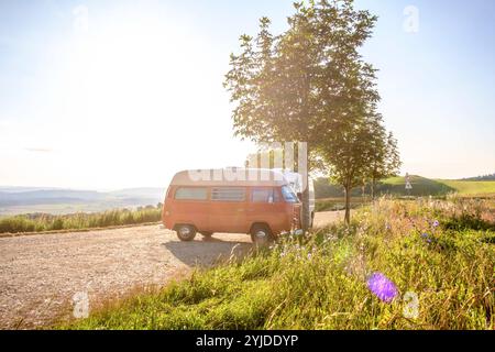 Ein Orange-weisser VW Bus Bulli steht auf einem Schotterparkplatz an einem Sommermorgen, Baden Württemberg, Deutschland. Ein Bulli steht in der Landschaft *** ein orange-weißer VW-Bus steht an einem Sommermorgen auf einem Schotterparkplatz, Baden-Württemberg, Deutschland Ein Van steht in der Landschaft Stockfoto