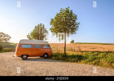 Ein Orange-weisser VW Bus Bulli steht auf einem Schotterparkplatz an einem Sommermorgen, Baden Württemberg, Deutschland. Ein Bulli steht in der Landschaft *** ein orange-weißer VW-Bus steht an einem Sommermorgen auf einem Schotterparkplatz, Baden-Württemberg, Deutschland Ein Van steht in der Landschaft Stockfoto