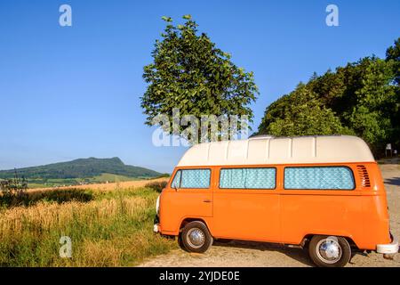 Ein Orange-weisser VW Bus Bulli steht auf einem Schotterparkplatz an einem Sommermorgen, Baden Württemberg, Deutschland. Ein Bulli steht in der Landschaft *** ein orange-weißer VW-Bus steht an einem Sommermorgen auf einem Schotterparkplatz, Baden-Württemberg, Deutschland Ein Van steht in der Landschaft Stockfoto