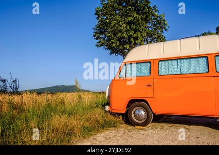 Ein Orange-weisser VW Bus Bulli steht auf einem Schotterparkplatz an einem Sommermorgen, Baden Württemberg, Deutschland. Ein Bulli steht in der Landschaft *** ein orange-weißer VW-Bus steht an einem Sommermorgen auf einem Schotterparkplatz, Baden-Württemberg, Deutschland Ein Van steht in der Landschaft Stockfoto