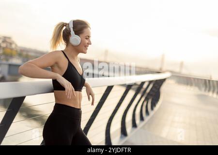 Während die Sonne über dem Wasser untergeht, lehnt sich eine junge Frau an ein Geländer und genießt ihre Lieblingsmusik über Kopfhörer, um Ruhe und Frieden zu genießen Stockfoto