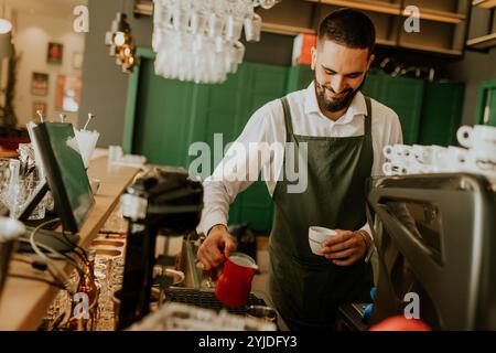 In einem warmen und einladenden Café gießt ein engagierter Barista fachmännisch Milch in eine Kaffeetasse und sorgt so für ein angenehmes Erlebnis für Gäste. Stockfoto