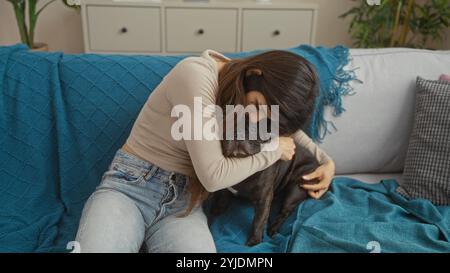 Eine junge hispanische Frau umarmt ihre französische Bulldogge auf einer gemütlichen Couch in einer modernen Wohnzimmeratmosphäre und bietet eine warme und liebevolle Atmosphäre. Stockfoto