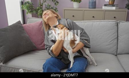 Eine reife blonde Frau lacht und umarmt ihren kleinen Hund, während sie auf einer Couch in einem gemütlichen Wohnzimmer sitzt, das mit Pflanzen und Kissen dekoriert ist. Stockfoto