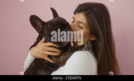 Eine junge hispanische Frau umarmt liebevoll ihr französisches Bulldogge-Haustier, isoliert vor einer rosa Hintergrundwand. Stockfoto