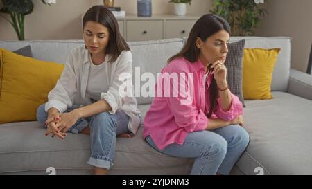 Zwei Frauen sitzen zusammen auf einer Couch in einem Wohnzimmer, erscheinen nachdenklich und beschäftigt mit verschiedenen Emotionen. Stockfoto