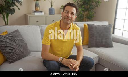 Hispanischer Mann, der in einem modernen Wohnzimmer sitzt, ein gelbes Hemd und Jeans trägt, mit Pflanzen und Kissen, die eine entspannte und nachdenkliche Demenz ausstrahlen Stockfoto