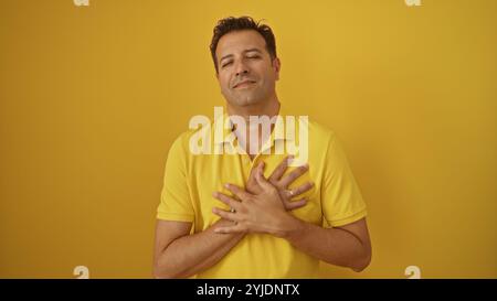 Ein hübscher hispanischer Mann posiert vor einem leuchtend gelben Hintergrund mit einem ruhigen Lächeln und überkreuzten Händen über der Brust mit einem passenden gelben Polo s Stockfoto