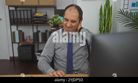 hispanischer Mann mittleren Alters im Büro, der Nackenschmerzen hat, während er an einem Schreibtisch sitzt, umgeben von Büromaterialien und Papierkram, was auf den Arbeitsplatz hinweist d Stockfoto