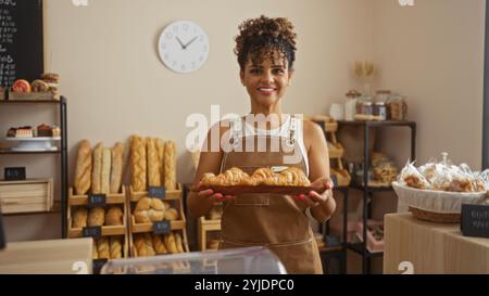 Junge Frau in einer Bäckerei, die Croissants hält, mit Brotregalen im Hintergrund und einer Uhr an der Wand, die selbstbewusst in die Kamera lächelt Stockfoto