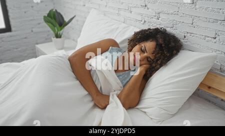 Schöne Frau mit lockigen brünetten Haaren, die friedlich in einem weißen Bett in einem gemütlichen Schlafzimmer mit einer Pflanze auf dem Nachttisch schläft Stockfoto