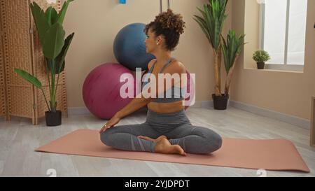 Frau übt Yoga in einem Wellness-Center, umgeben von Pflanzen und Übungsbällen, und demonstriert eine sitzende Twist-Pose auf einer Yoga-Matte in einem ruhigen Spa-Set Stockfoto