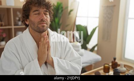 Junger Mann, der einen ruhigen Moment in einem Spa-Center genießt, in einem Bademantel gekleidet mit Händen in einer Gebetsposition, umgeben von beruhigendem und ruhigem Wellness Stockfoto