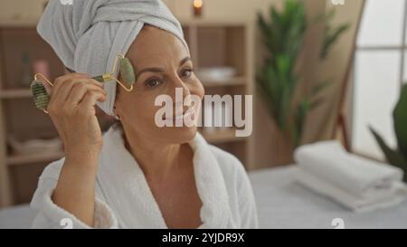 Eine hispanische Frau mittleren Alters in einem Spa-Raum, die eine Jaderolle auf ihr Gesicht aufträgt, einen weißen Bademantel mit ruhigem Ausdruck trägt, umgeben von einem ruhigen W Stockfoto