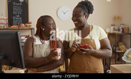 Zwei glückliche afroamerikanische Frauen stehen in einer Bäckerei, eine mit Telefon und die andere mit einer Kaffeetasse, die Schürzen trägt und sich anlächelt. Stockfoto