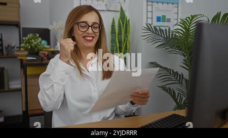 Frau feiert Erfolg im modernen Büro mit Computer und Dokumenten, zeigt blondes Haar, Brille und ein weißes Hemd Stockfoto