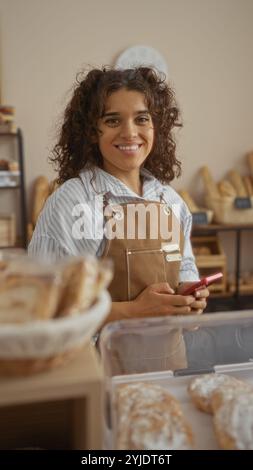 Junge, lächelnde Frau in brauner Schürze, die in einer Bäckerei steht und ein pinkfarbenes Telefon mit verschiedenen Broten im Vordergrund und Regalen im Hintergrund hält Stockfoto