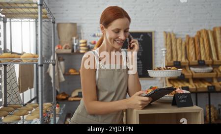 Rothaarige Frau in einer Bäckerei, die ihr Tablet überprüft, während sie am Telefon telefoniert, umgeben von verschiedenen Backwaren und Gebäck in einem gemütlichen Innengeschäft Stockfoto