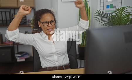 Begeisterte Frau, die im Büro mit Computer feiert, Brille und weißem Hemd trägt und Erfolge am Arbeitsplatz zeigt. Stockfoto