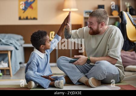 Kleiner lächelnder Schwarzer Junge, der dem fröhlichen Adoptivvater hohe fünf schenkt, während er den Sieg beim Handy-Spiel feiert, der auf dem Boden im Kinderzimmer sitzt Stockfoto